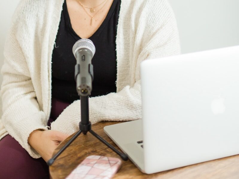 How to Start a Podcast with No Audience Feature Image, woman wearing a white cardigan over a black tee recording her podcast episode.